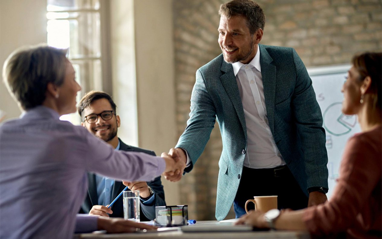 Business professionals shaking hands during a successful meeting, with colleagues smiling and a presentation board in the background.