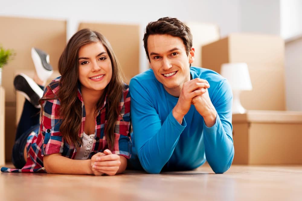 Smiling couple in casual clothes relaxing in their new home with cardboard boxes in the background, symbolizing first-time home buyers in Ontario.