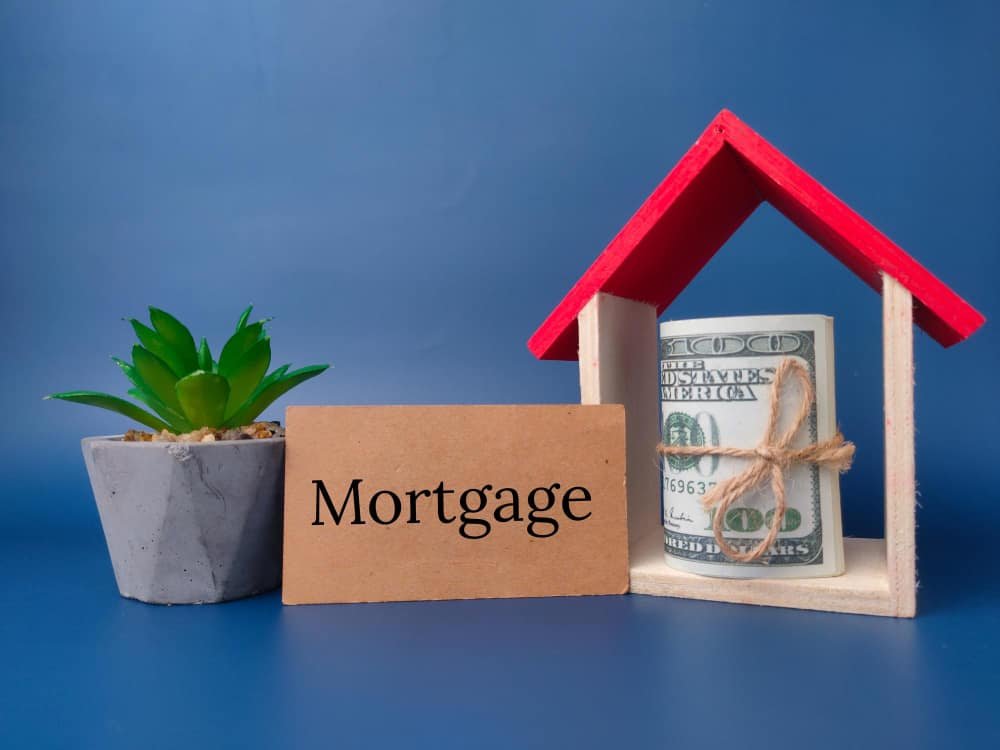 Rolled cash under a toy house roof beside a 'Mortgage' sign, symbolizing deposit requirements for first-time home buyer programs.