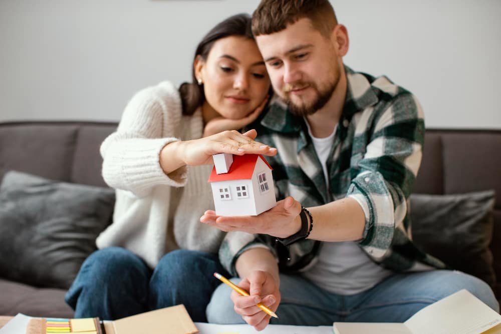 Young couple holding model house while planning finances, symbolizing first-time home buying and mortgage decisions.
