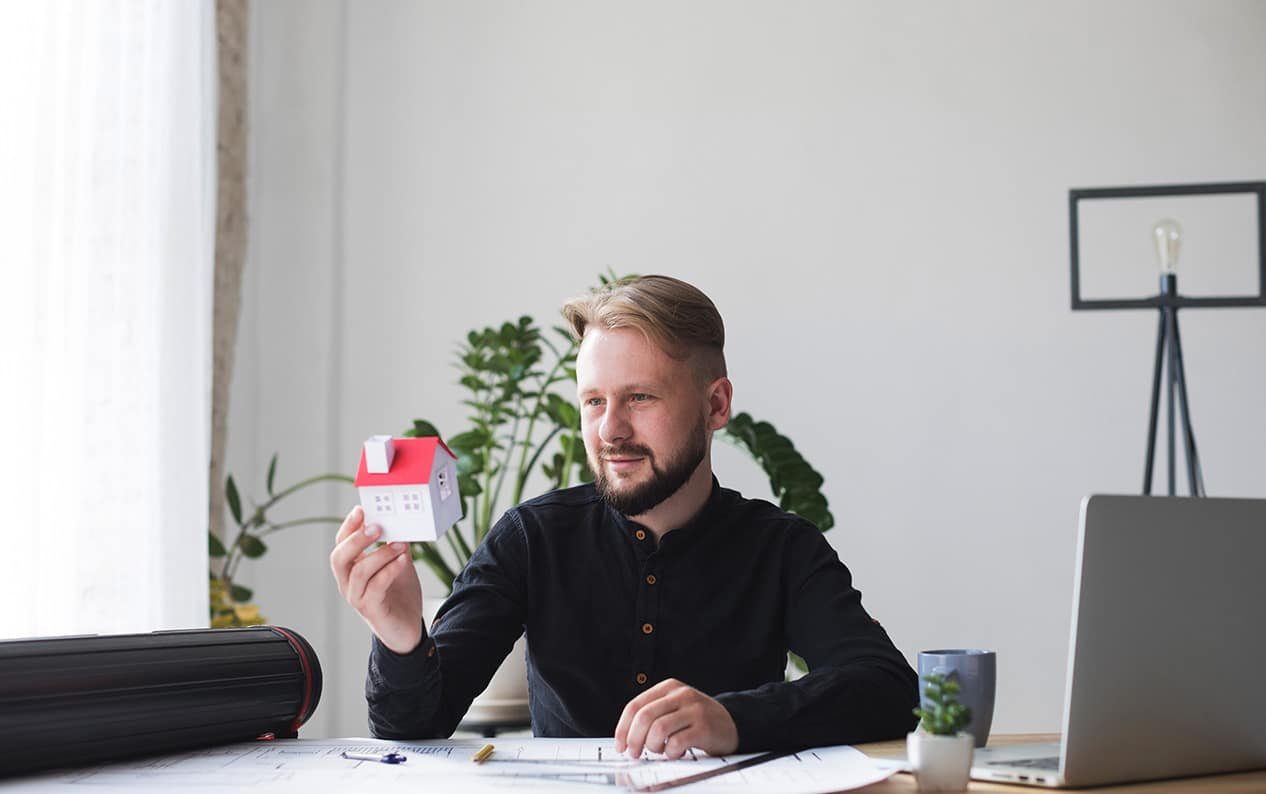 A man, possibly self-employed in Ontario, is sitting at a desk with blueprints and a model house, perhaps considering a mortgage for a new property.