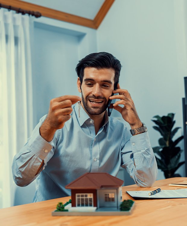Smiling man holding house key and talking on phone, with model house on desk, symbolizing smart home equity planning