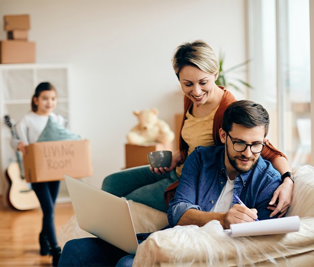 Smiling couple reviewing notes with a laptop on a couch while their child carries a moving box in the background, symbolizing planning for a second mortgage